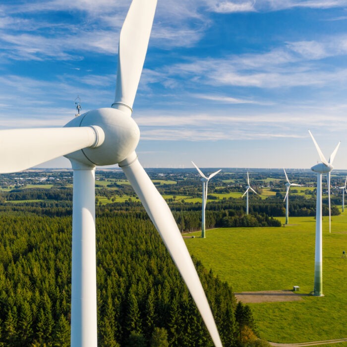 A close-up of a white wind turbine in a large, green field with a forest behind it.