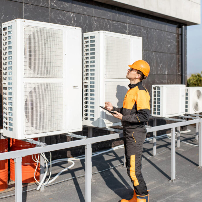 An engineer dressed in orange and black overalls and a yellow hard hat checks on a heat pump system with a tablet.