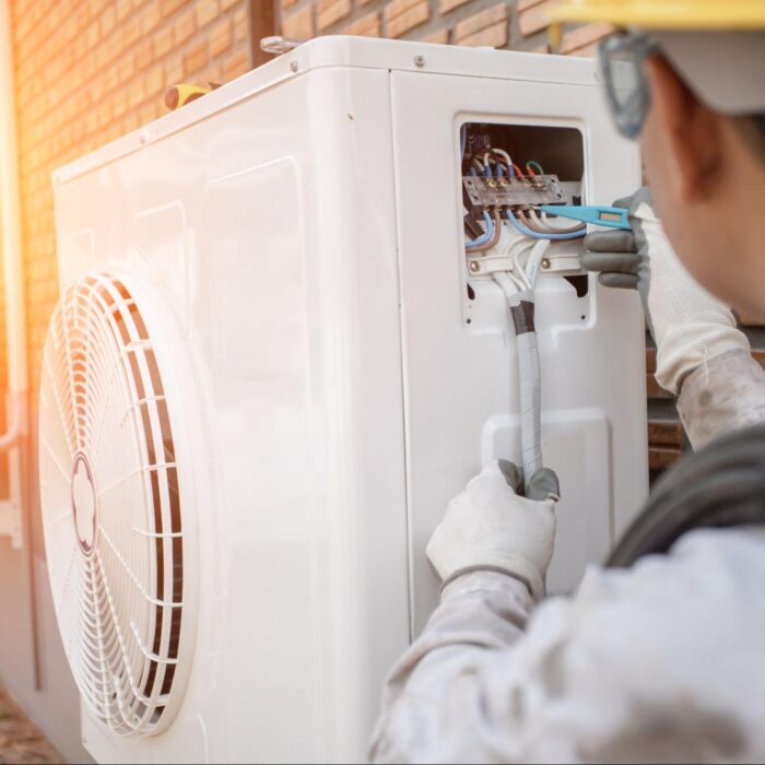 An engineer dressed in white overalls and a yellow hard hat instals a new heat pump system to a property.